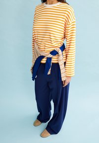 Langärmliges, gelb-weiß gestreiftes Hemd mit einem beigen und blauen Pullover, der um die Taille gebunden ist; kombiniert mit navyblauen, weit geschnittenen Hosen und beigen Ballerinas.