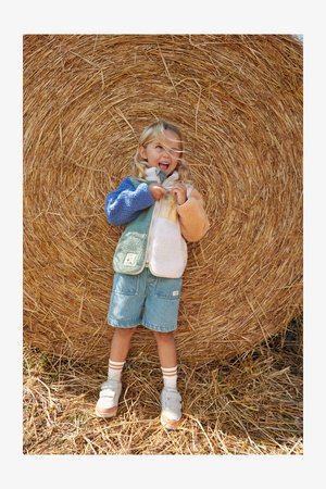 Színes, többrészletű polárdzseki, cipzáros elülső résszel, zöld, kék, narancs és krémszínű részekkel. Farmer rövidnadrággal és fehér sportcipőkkel viselve.