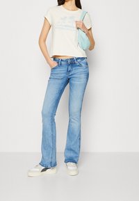 Ljusblå utsvängda jeans med subtil blekning, kombinerat med en krämfärgad kort t-shirt som har en pastellgrafik och en ljusblå axelväska.