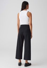 Haut sans manches blanc associé à un pantalon large noir taille haute. Le pantalon possède deux poches arrière et une texture lisse. Chaussures noires à enfiler.
