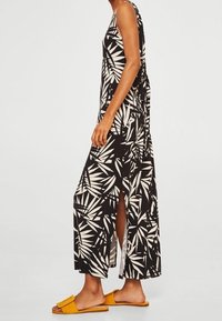 Schwarzes Maxikleid mit weißem Blattmuster, ärmelloses Design, seitliche Schlitze für mehr Bewegungsfreiheit, kombiniert mit gelben Rutschsandalen.