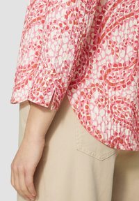 Arm en hand van een persoon die beige broek draagt en een langemouwen shirt met een roze en witte bloemenprint en een split in de manchet.