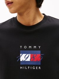 Černé bavlněné tričko s grafickým logem, na kterém jsou bílé nápisy "TOMMY," "1985" a "HILFIGER", doplněné modrými a červenými obdélníky.