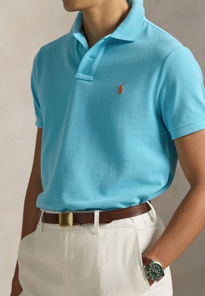 Ανοιχτό γαλάζιο πόλο με κοντά μανίκια, διακριτή υφή, πορτοκαλί λογότυπο στο στήθος, συνδυασμένο με ανοιχτόχρωμο παντελόνι και καφέ ζώνη.