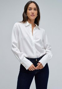 Mujer vestida con una camisa blanca de botones de mangas largas y jeans de color azul oscuro, de pie contra un fondo gris claro liso.