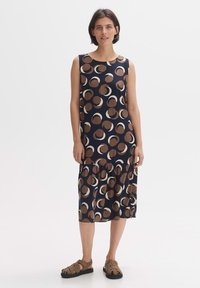 Sleeveless navy dress with a ruffled hem, featuring a cream and brown circular pattern, made of lightweight fabric. Worn with brown sandals.