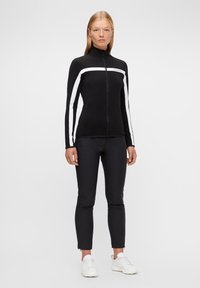 Svart idrottsjacka med vita ränder, hög krage, full dragkedja. Parat med svarta tighta byxor och vita sneakers. Slät textur.