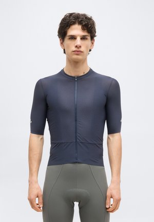 Młody mężczyzna stoi, ubrany w dopasowaną granatową, krótką koszulkę kolarską z zamkiem z przodu oraz szare, wyściełane spodenki kolarskie.