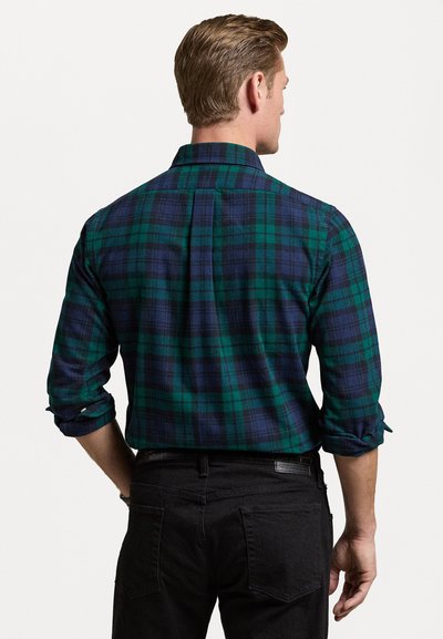 Groene en marineblauwe geruite blouse met opgerolde mouwen en een rugplooitje, gemaakt van zachte stof, gecombineerd met zwarte broek, bekeken vanuit de achterkant.