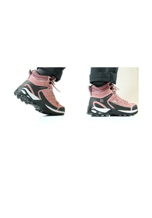 Paire de chaussures de randonnée roses et noires portées avec un pantalon noir, vues de côté et par en dessous sur fond blanc.