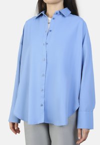 Camisa azul de manga larga en una tela suave con cuello puntiagudo, frente abotonado y corte holgado, combinada con pantalones grises.