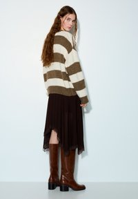 Pull en maille rayé avec des couleurs crème et olive, associé à une jupe longue marron fluide avec un bord en dentelle, et des bottes en cuir marron montant jusqu'aux genoux.