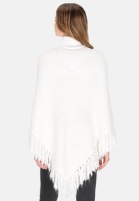 Nainen, jolla on valkoinen fringed-poncho valkoisen poolopaidan päällä, seisoo selkä kameraan päin vaaleanharmaata taustaa vasten.