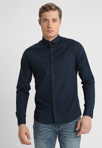 Dunkelblaues, langärmliges Button-Down-Hemd mit Kragen und Vorderfrontverschluss, aus glattem Stoff. Getragen mit hellblauen Jeans.