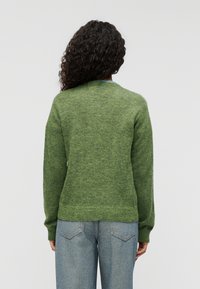 Πράσινο πλεκτό πουλόβερ με μαλακή υφή, ριμπ μανσέτες και στρογγυλή λαιμόκοψη, συνδυασμένο με ανοιχτό μπλε τζιν παντελόνι.