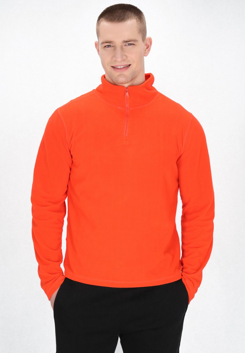 Orange fleece pullover jakke med høj krave og kvart lynlås. Har lange ærmer og en glat tekstur. Sammen med mørke bukser.