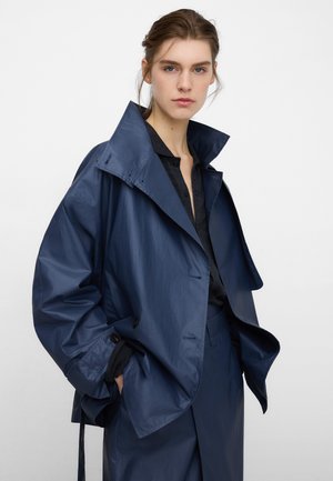 Giacca leggera blu navy con collo alto, dettagli con bottoni e vestibilità rilassata. Tessuto strutturato, maniche lunghe e tasche laterali.