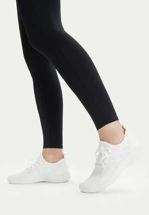 Nogi w czarnych leginsach noszące białe dziergane trampki na sznurowadła, stojące na jednolitym białym tle.