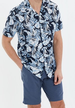 Homme portant une chemise bleu marine à manches courtes avec un imprimé feuilles tropicales bleu clair et un short bleu uni, les mains dans les poches.