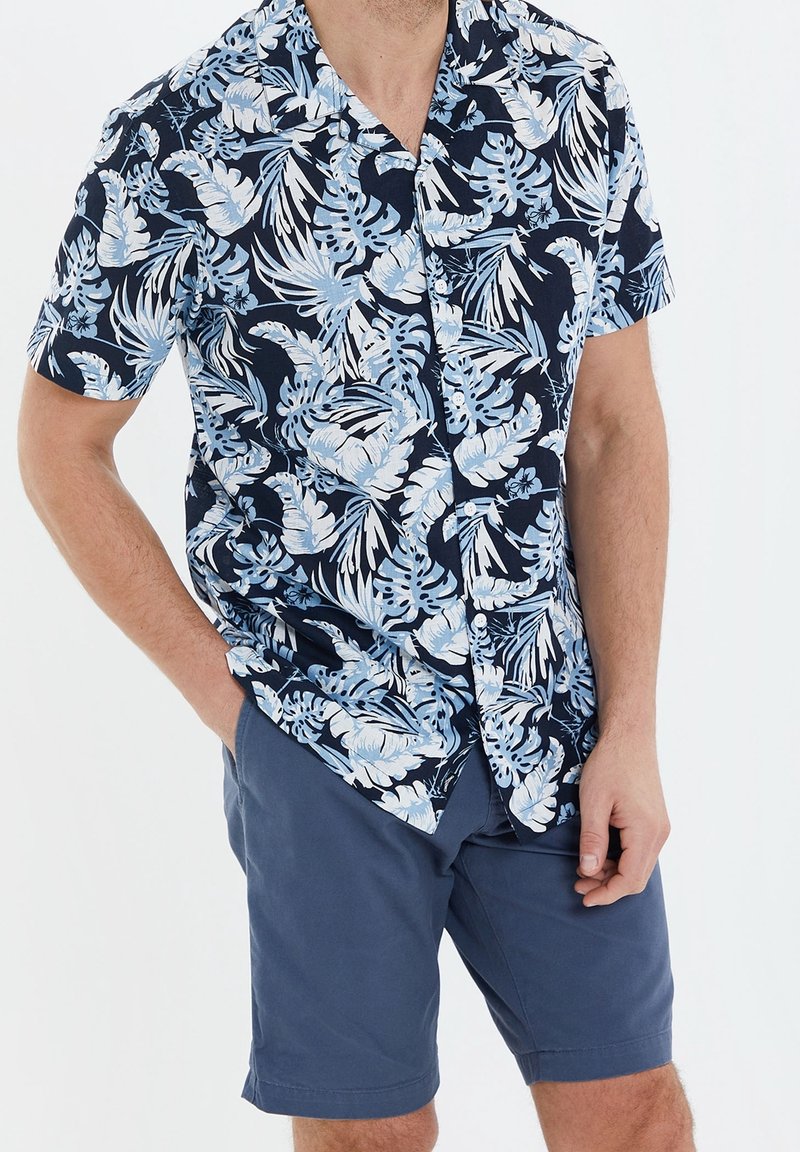 Homme portant une chemise bleu marine à manches courtes avec un imprimé feuilles tropicales bleu clair et un short bleu uni, les mains dans les poches.