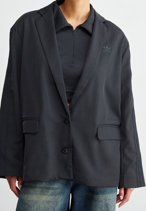 Osoba nosząca czarną oversize marynarkę na czarnym topie Adidas z zamkiem i niebieskich dżinsach na jednolitym tle.