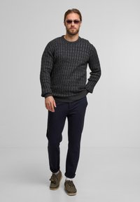 Dunkelgrauer gestrickter Pullover mit strukturiertem Muster, kombiniert mit dunkelblauen Hosen und braunen Mokassins. Sonnenbrillen runden den Look ab.
