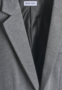 Sivi blejzer s glatkom teksturom i oštrim reverima. Unutrašnjost ima tamniju podlogu. Oznaka prikazuje "ANTONY MORATO."