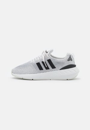 Hvid atletiksneaker med sorte striber, strikket overdel, hvide snørebånd og tyk hvid sål på en ensfarvet lys baggrund.