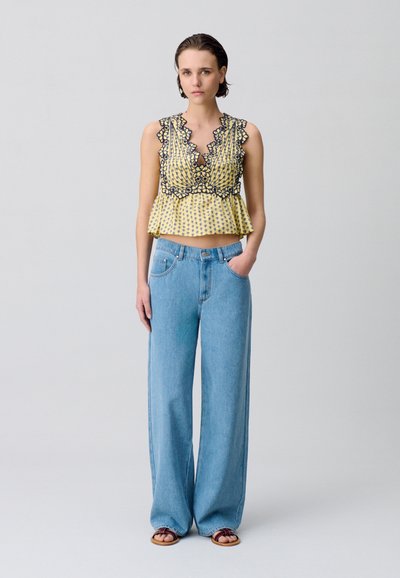 Top giallo a peplo con bordo in pizzo nero e design plissettato; abbinato a jeans a vita alta, larghi e di colore blu chiaro.