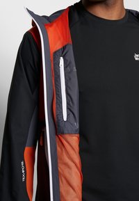 Zwarte sportieve T-shirt onder een ritsjack met grijze en oranje accenten; bevat een mesh voering en een ritszak op de borst.