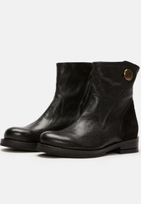 Botas de cuero negro hasta el tobillo con un acabado suave, punta redonda, costuras laterales y un botón decorativo de latón en el lado exterior. Suela plana.