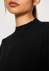 Black knitted pullover with a high neckline. Soft texture, ribbed detail on collar, and fitted style. Close-up view focusing on the neckline.