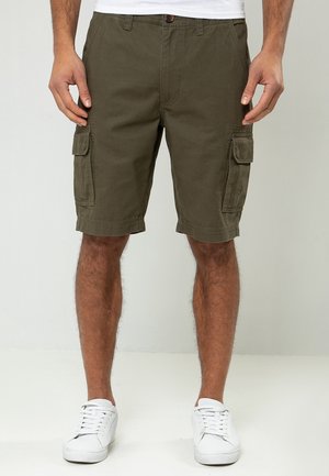 Mand iført olivengrønne cargoshorts med sidelommer og hvide sneakers, stående foran en ensfarvet baggrund.