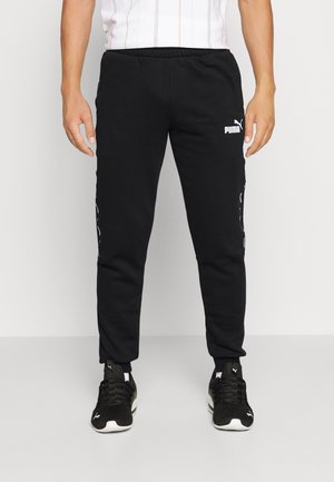 Mann trägt schwarze Puma-Sweathosen und schwarze Puma-Sneaker und steht vor einem einfarbigen Hintergrund.