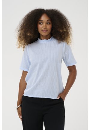 Žena s kovrčavom kosom nosi svijetloplavu prugastu majicu kratkih rukava i crne hlače, stoji s jednom rukom u džepu nasuprot jednostavnoj pozadini.