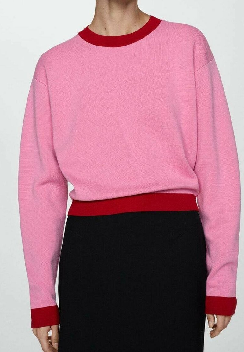 Personne portant un pull rose avec un col, des poignets et une ceinture rouges, associé à une jupe ou un pantalon noir. Haut du corps montré, visage non visible.