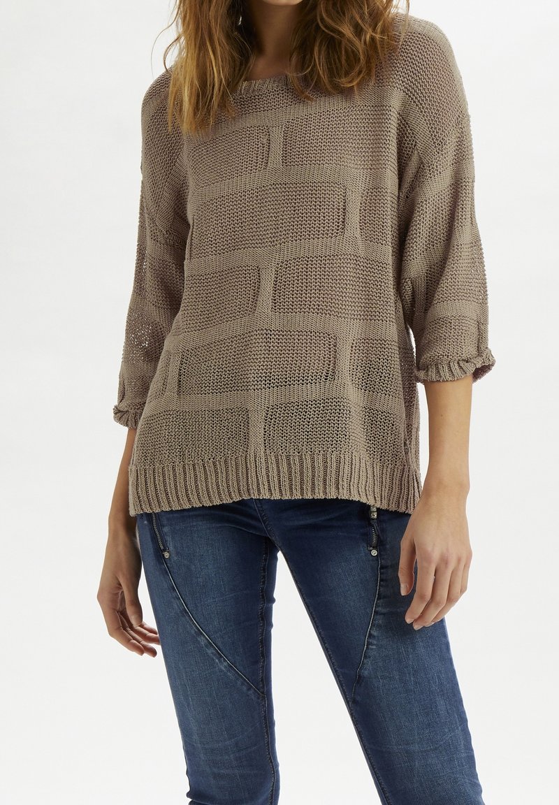Femme portant un pull ample en tricot taupe avec un motif rectangulaire texturé et un jean bleu avec des détails de couture diagonale.