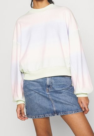 Personne portant un sweat tie-dye pastel clair avec des manches bouffantes et une mini-jupe en jean bleu, debout devant un fond uni.