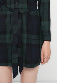 Robe chemise à carreaux vert et noir avec des manches longues, fermeture par boutons et une ceinture en tissu. Texture douce et coupe droite au niveau de l'ourlet.