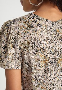 Blouse à motif floral avec manches courtes bouffantes, palette de couleurs beige et noire avec accents dorés, et fermeture dos en trou de serrure.