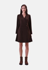 Bruine wikkeljurk met lange mouwen, een aansluitende taille en een geplooide rok. Kenmerkend is een knoopaccent. Model draagt bruine enkellaarsjes.