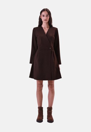 Braunes Wickelkleid mit langen Ärmeln, tailliertem Design und gefaltetem Rock. Besitzt eine Knopfakzentuierung. Das Model trägt braune Knöchelstiefel.