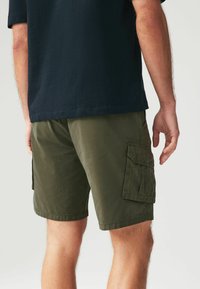 Șorturi cargo verzi cu buzunare laterale, fabricate dintr-un material durabil. Au o croială lejeră și tivuri, fiind asociate cu un tricou închis la culoare.