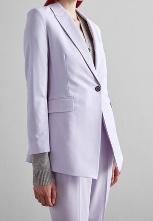 Femme portant un blazer sur mesure lavande clair avec un bouton noir, combiné à un pantalon assorti et un haut gris à manches longues en dessous.