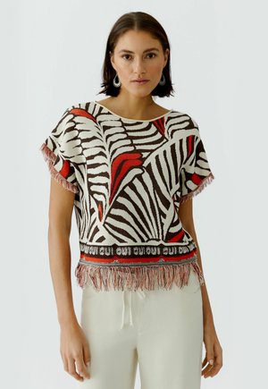 Frau trägt ein kurzärmeliges, mit Fransen versehenes Oberteil mit abstrakten roten, schwarzen und weißen Mustern, kombiniert mit hellen Hosen und hängenden Ohrringen.