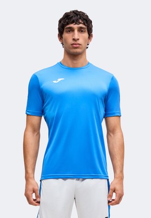Homme aux cheveux courts et bouclés portant un t-shirt de sport bleu uni et un short blanc, debout sur un fond gris clair.