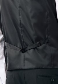 Gilet en satin noir à la texture lisse, avec un dos élégant, une boucle réglable et deux poches latérales sur un costume sur mesure.