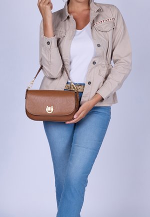 Mujer sosteniendo un bolso de piel marrón con cierre dorado, lleva chaqueta beige, camiseta blanca, vaqueros azules y cinturón beige sobre fondo liso.