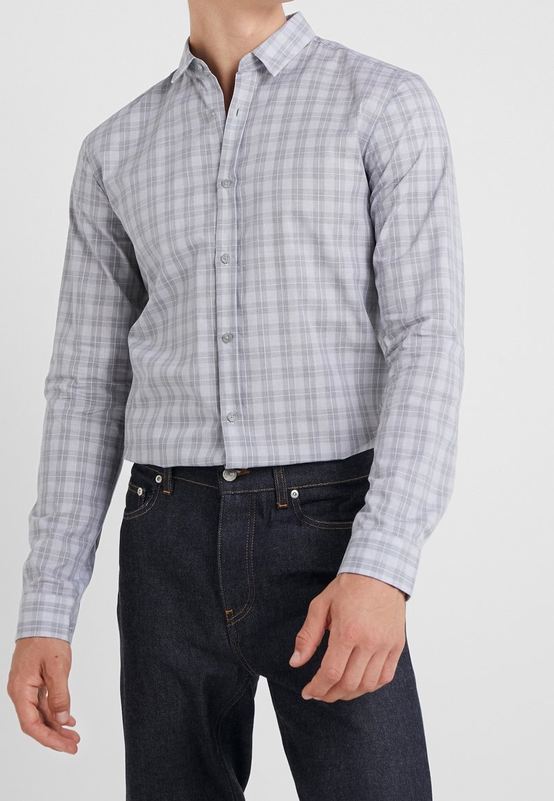 Homme portant une chemise boutonnée à carreaux gris clair et un jean bleu foncé, debout devant un fond uni.
