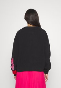 Svart stickad tröja med lös passform och ribbad textur, prydd med rosa blommig broderi på ärmarna; kombinerat med en rosa plisserad kjol.